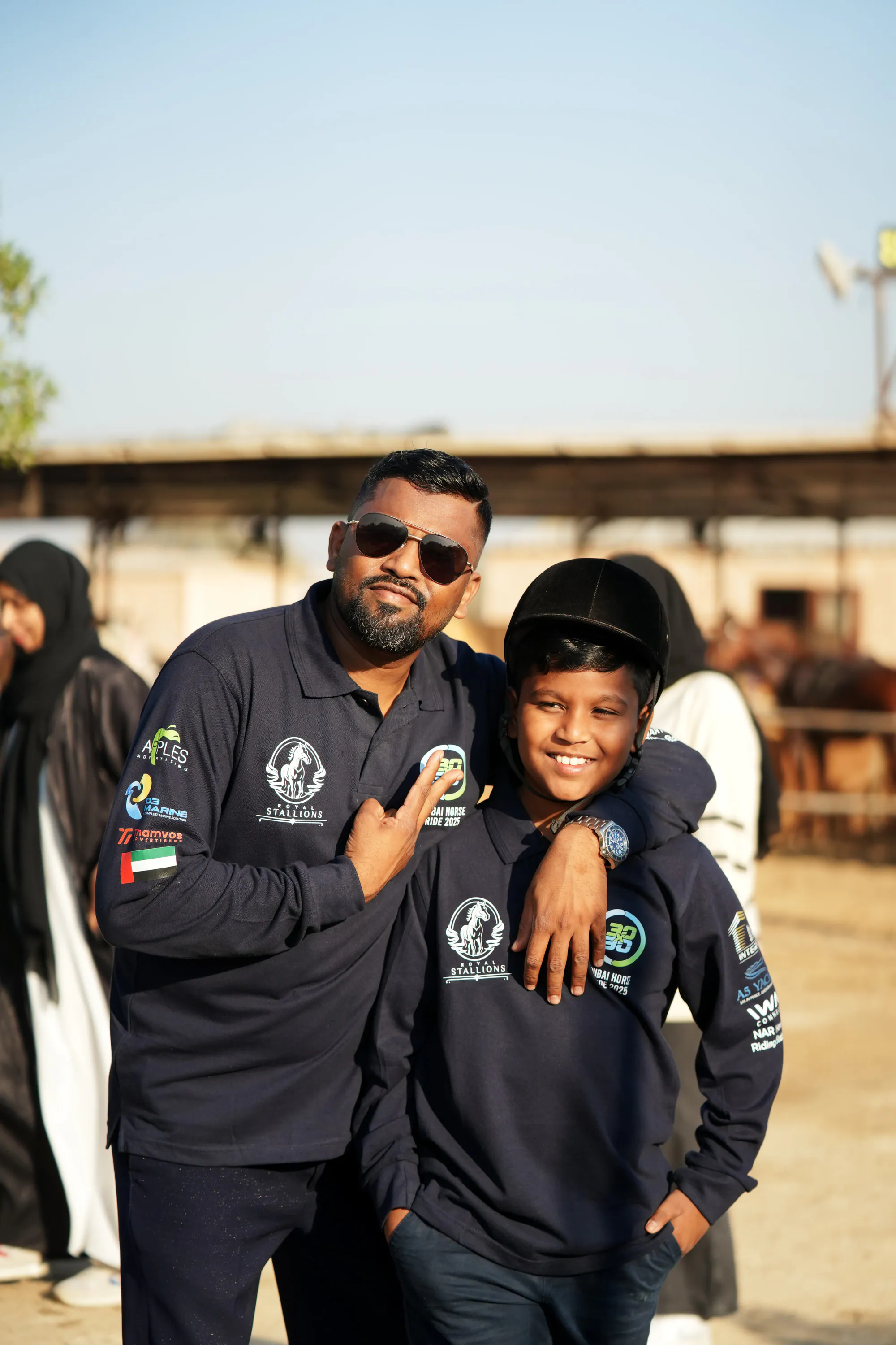 A father and son in Royal Stallions jackets smiling at the stables