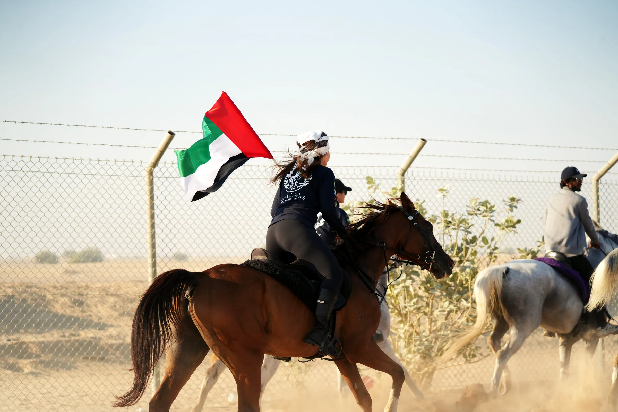 A rider cantering a chestnut horse while holding a large UAE flag